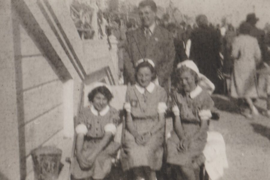Blurred black and white photograph showing three Nursing Cadets seated in deckchairs at the bottom of a staircase onto a busy promenade. Behind them stands a man in a suit. The Cadets all wear grey knee length dresses with white nursing caps and white cuffs at their elbows.  