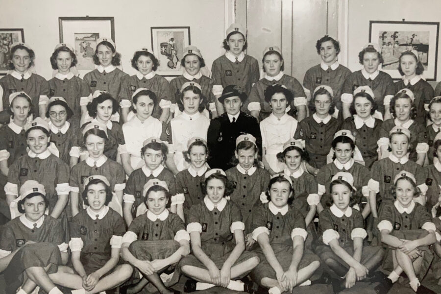 Black and white photograph of 40 young girls seated in four rows. They all wear nurse uniforms featuring white nursing caps and white cuffs. On the walls behind them are four framed pictures