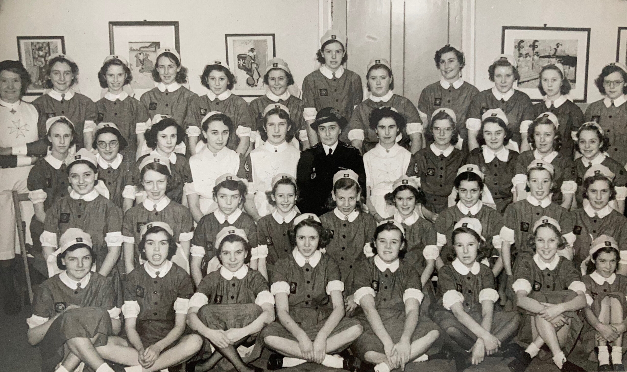 Black and white photograph of 40 young girls seated in four rows. They all wear nurse uniforms featuring white nursing caps and white cuffs. On the walls behind them are four framed pictures