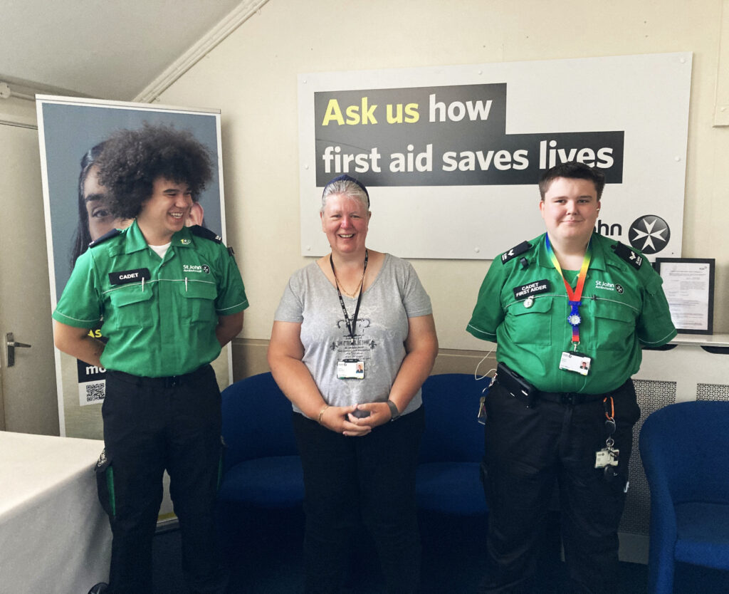Colour photograph of two Cadets standing either side of an older woman. Behind them is a blue sofa and a wall graphic that reads &apos;Ask Us How St John Saves Lives&apos;. They are all smiling.