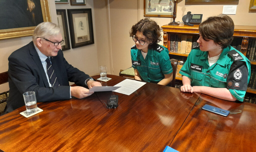 Colour photograph of an older man in a suit seated at a polished wooden table. Opposite him are two Cadets in green and black St John uniform. The man is showing them a document and pointing at something on it. There is a black recorder on the table between them.