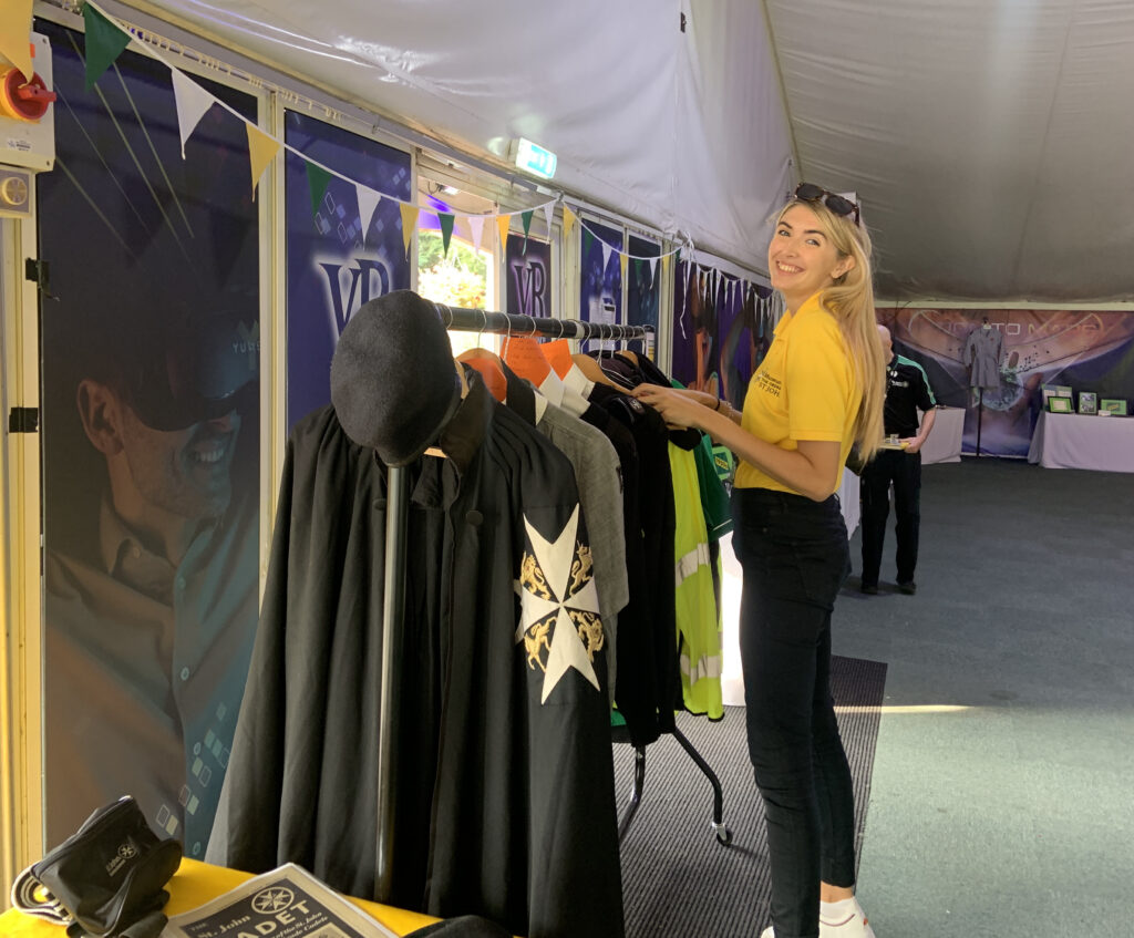 Colour photograph of a young woman in a yellow t-shirt and black trousers smiling at the camera whilst she reaches out to some uniform that is hanging on a clothes rail in front of her. The uniform items are mainly black and grey and are not clearly visible. The backdrop is the inside of a marquee.