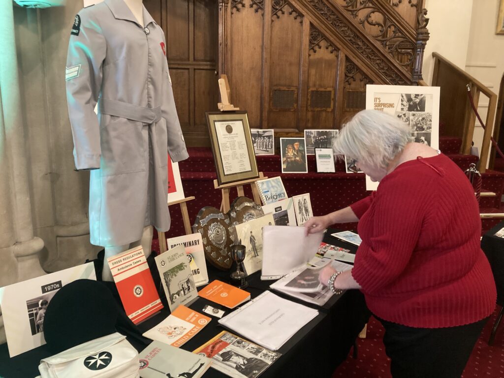 Colour photograph of a woman browsing some old photographs that are laid out on a table. The table is full of different types of artefact including a grey nursing dress on a mannequin, a white bag decorated with a black eight-pointed cross, shields, magazines and booklets.
