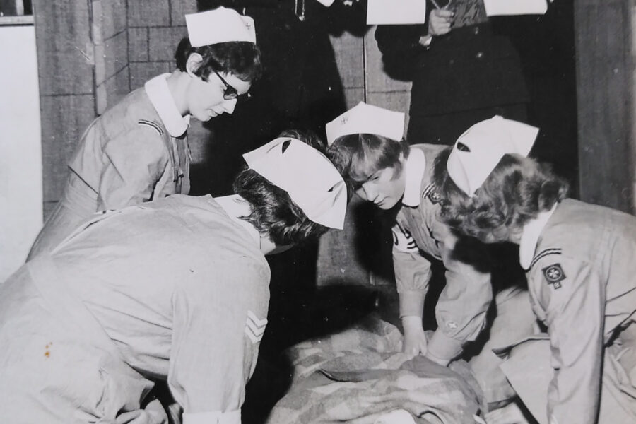 Black and white photograph of four female Cadets in uniform treating a person who is lying on the floor as a casulty simulation. Three adults in uniform are looking on and making notes on a clipboard.