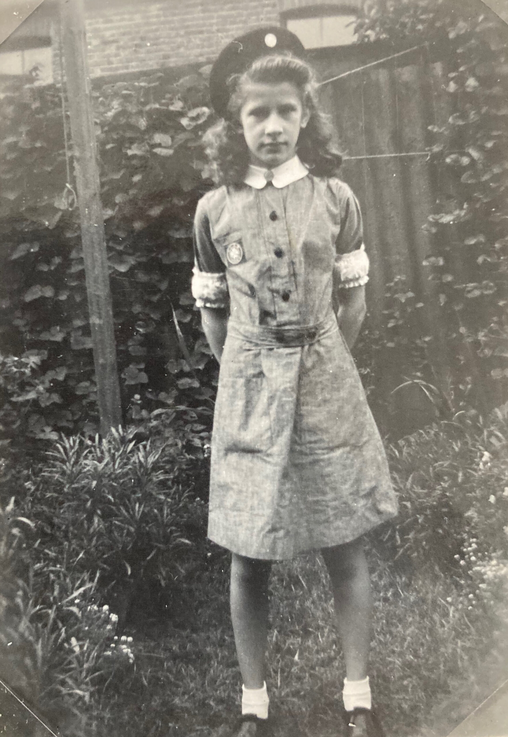 Black and white photograph of a young female Cadet in uniform. She wears a dress and a beret and is standing in a garden.
