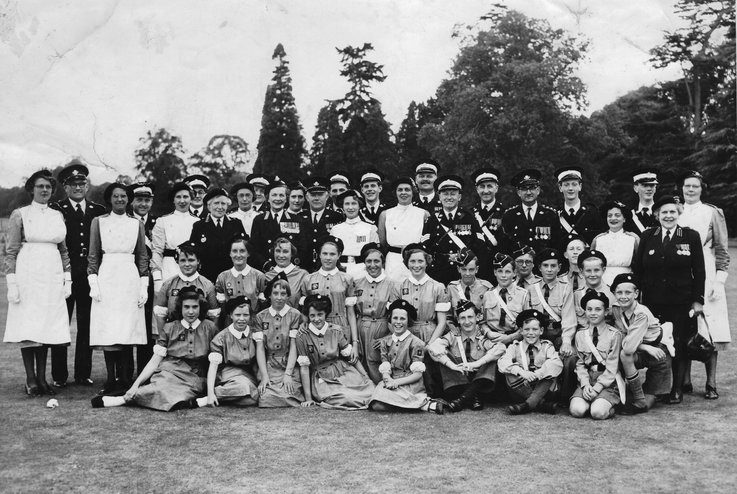 A black and white group photograph of the Winchester St John Ambulance Brigade Division. In the foreground seated on the floor are female Nursing Cadets wearing grey dresses and berets and male Ambulance Cadets wearing grey shirts with white haversacks worn across their bodies. Behind the Cadets stand adult nursing and ambulance members. The men are wearing St John Ambulance’s black and white number one uniform and the women are wearing knee length grey dresses with white aprons over the top. 