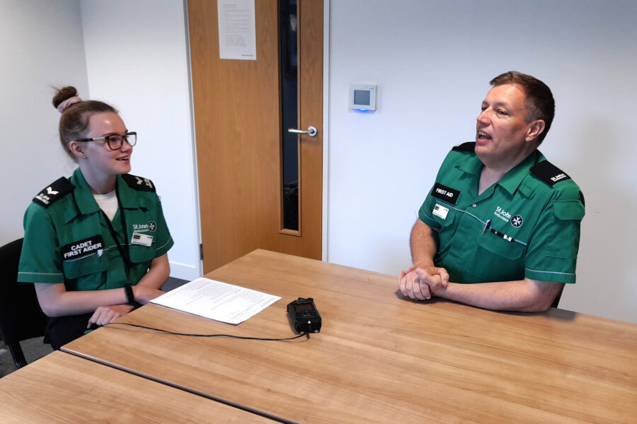 Colour photograph of a young female Cadet in green St John Ambulance uniform sitting at a wooden table with an adult male also in green uniform. Between them there is an audio recorder on the table.