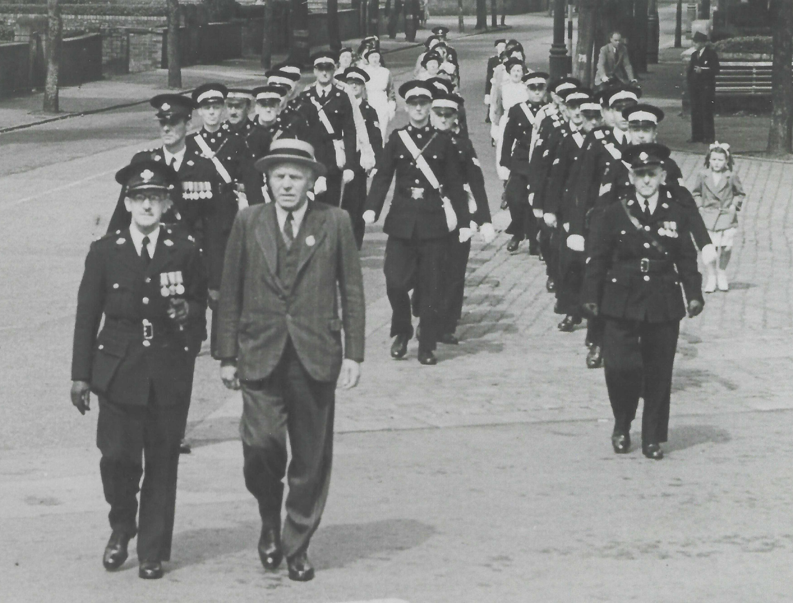 Black and white images of three rows of men in uniform who are walking in the direction of the photographer.  Behind them are three rows of nurses in uniform