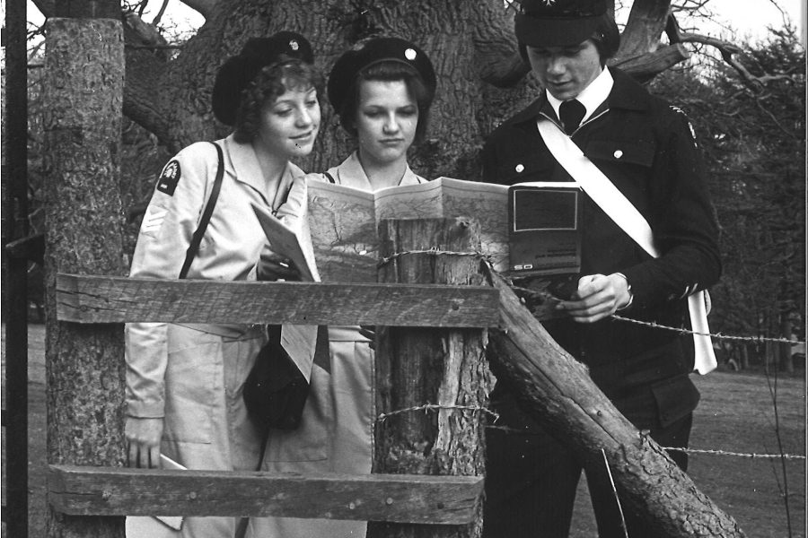 Black and white photograph of three people stood by a gate reading a map. There are two women in uniform and on the right a man in uniform. The girls wear white dresses and black hats. The man wears a black suit and black hat. He is carrying a small white bag across his body. Behind them is the trunk of a large tree.