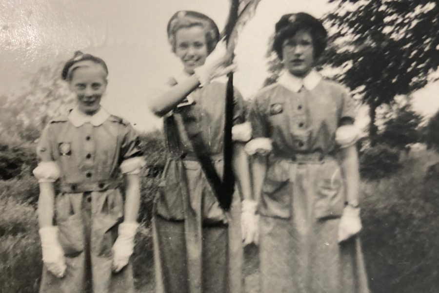 Black and white photograph of three female Cadets standing outside in a line facing the camera. The middle Cadet holds a flag on a pole.