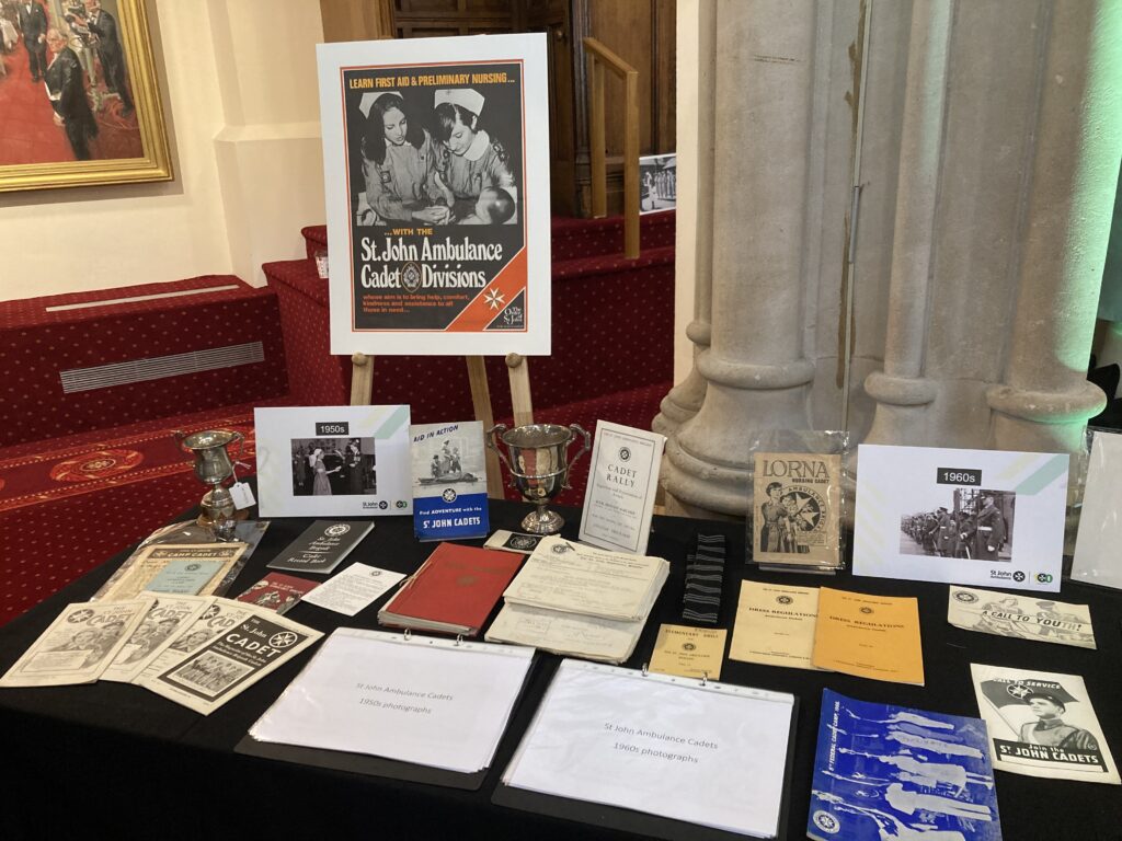 Colour photograph of a table covered with old photographs, silver trophies, magazines and a black and white striped tie. A poster entitled &apos;St John Ambulance Cadet Divisions&apos; with a black and white photograph of two girls in St John uniform washing a baby is displayed on a wooden easel behind the table.