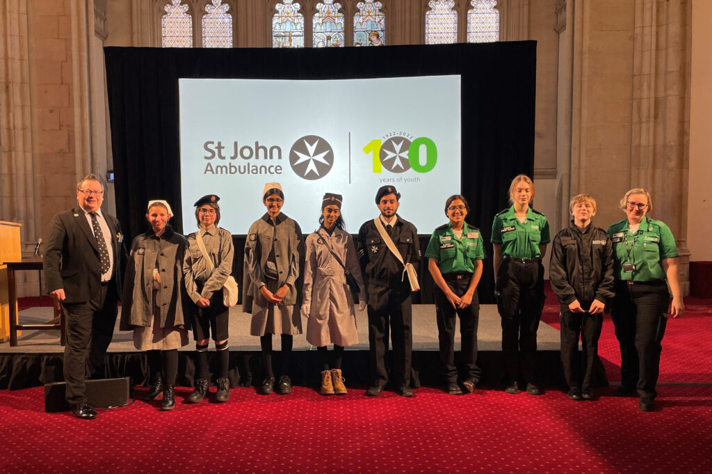 Colour photograph of two adults and 8 Cadets standing in front of a stage with a screen displaying the St John Ambulance Year of Youth logo. The Cadets are wearing different St John uniforms to illustrate how they have changed from the 1920s to the present day. The backdrop behind the stage is a grand setting with a high ceiling, stone walls and stained glass windows.