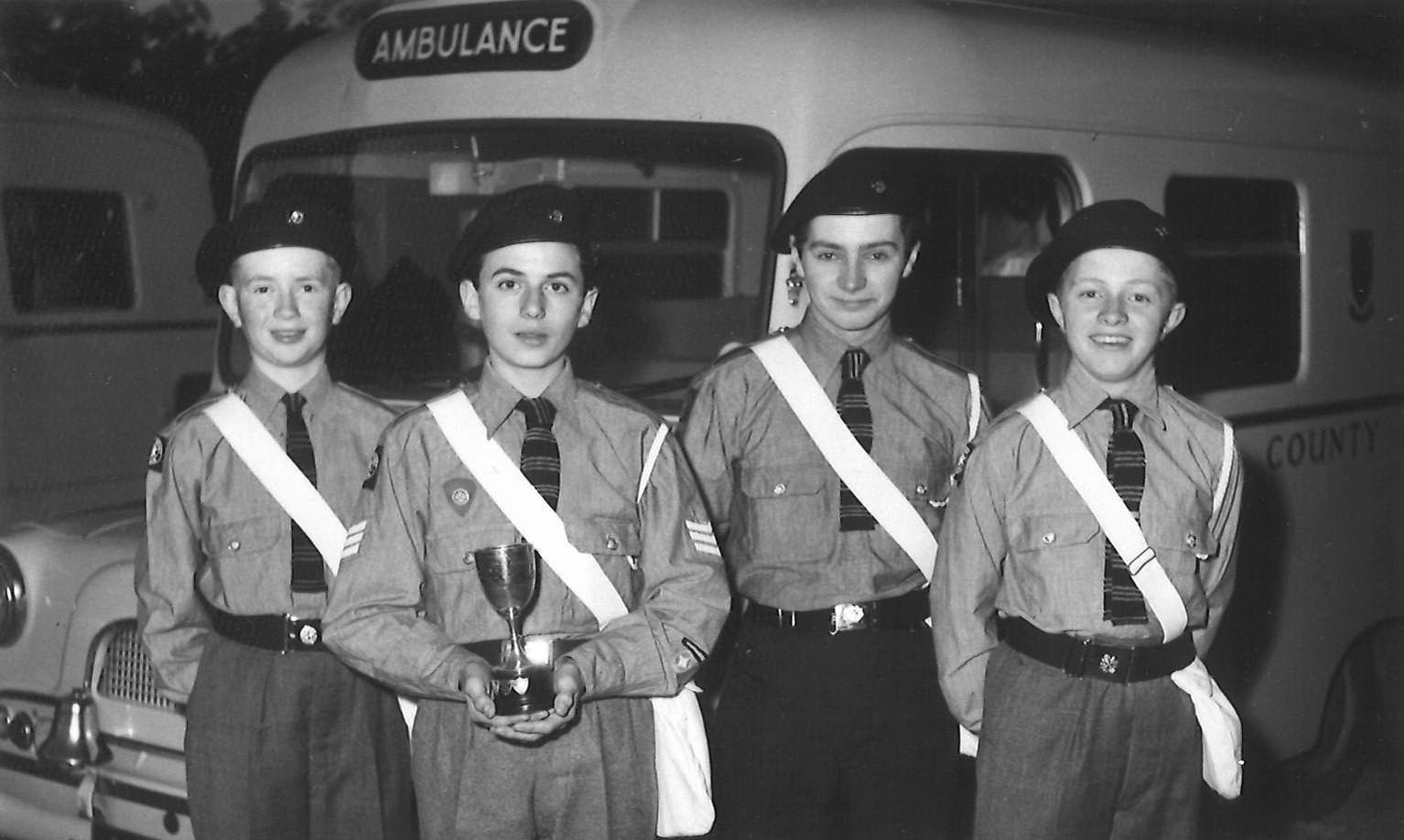 A black and white photograph showing four young St John Ambulance Cadets standing in front of an old ambulance. The Cadets are wearing berets on their heads, grey shirts, stripey ties, and a white haversack across their bodies. Their dark grey trousers are belted at the waist with a black belt and silver clasp. The Cadet second from left, holds a small trophy close to his body with both hands.