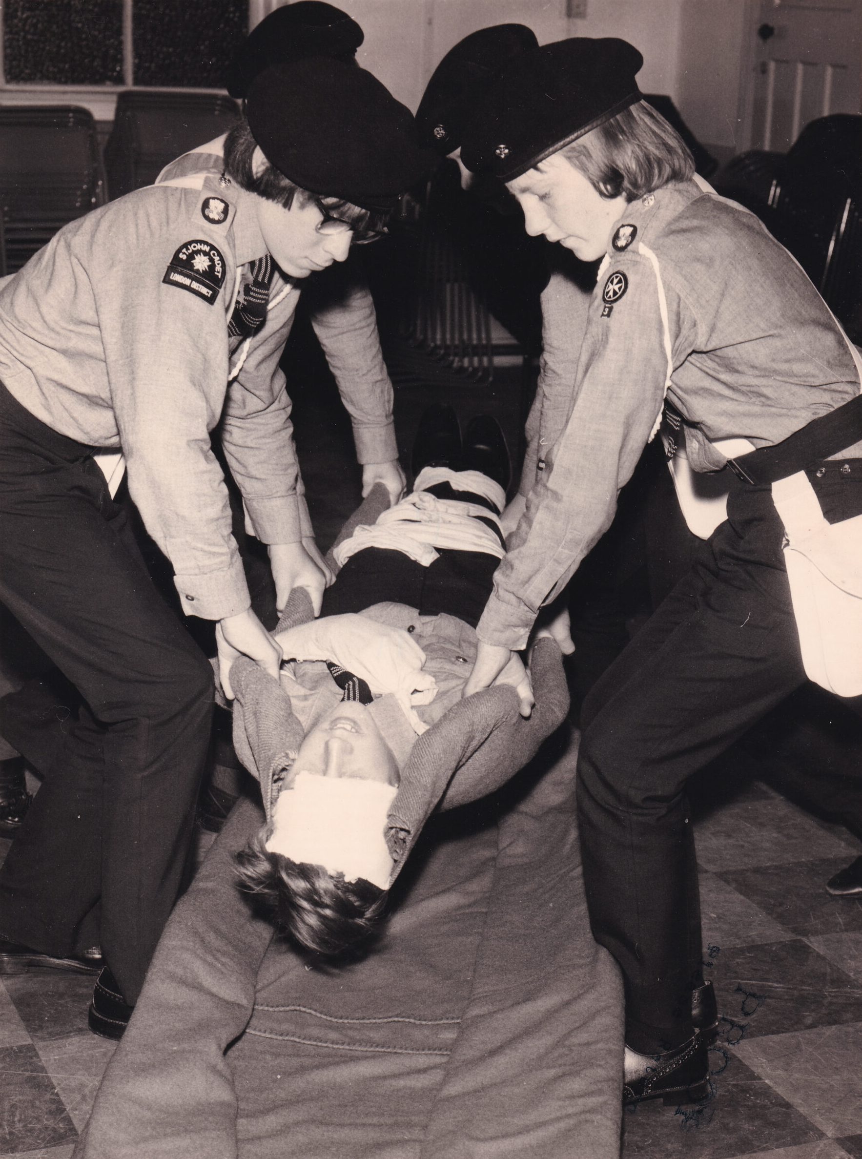 Black and white photograph of four Cadets in uniform treating a fifth Cadet who is lying on the floor as a casulty simulation. They are in the process of moving the lying person to a stretcher.