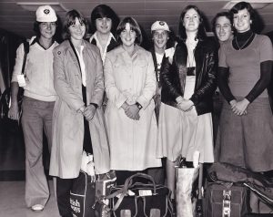 Black and white photograph of 8 teenagers standing in a row. Two wear St John Ambulance caps. They are smiling at the camera. At their feet are some bags.
