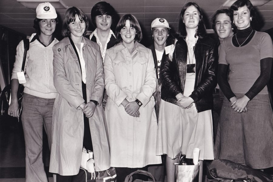Black and white photograph of 8 teenagers standing in a row. Two wear St John Ambulance caps. They are smiling at the camera. At their feet are some bags.
