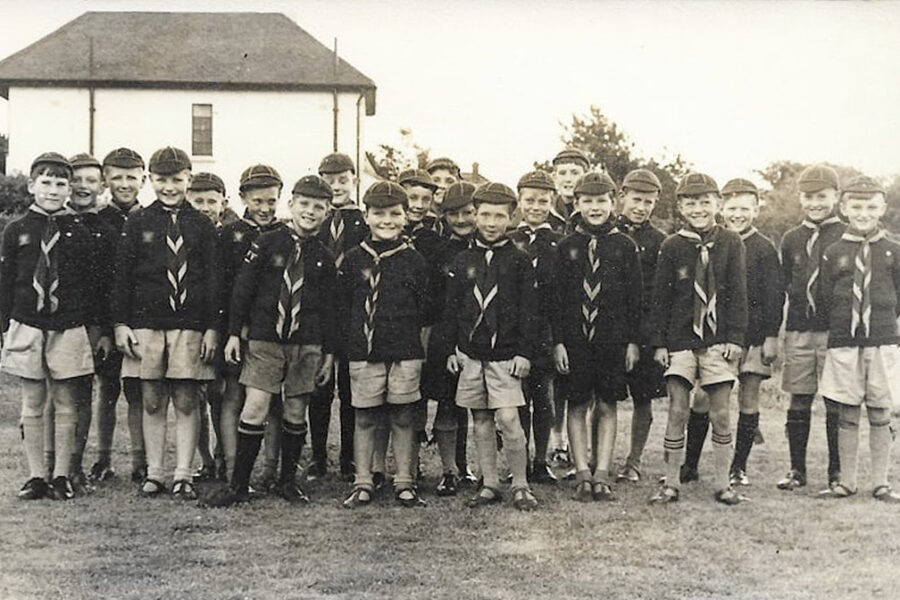 Black and white photograph of 21 young boys standing in a line facing the photographer. They all wear Cub Scout uniforms. Behind them is the side of a white building.