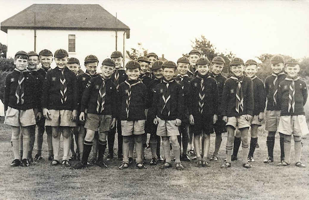 Black and white photograph of 21 young boys standing in a line facing the photographer. They all wear Cub Scout uniforms. Behind them is the side of a white building.