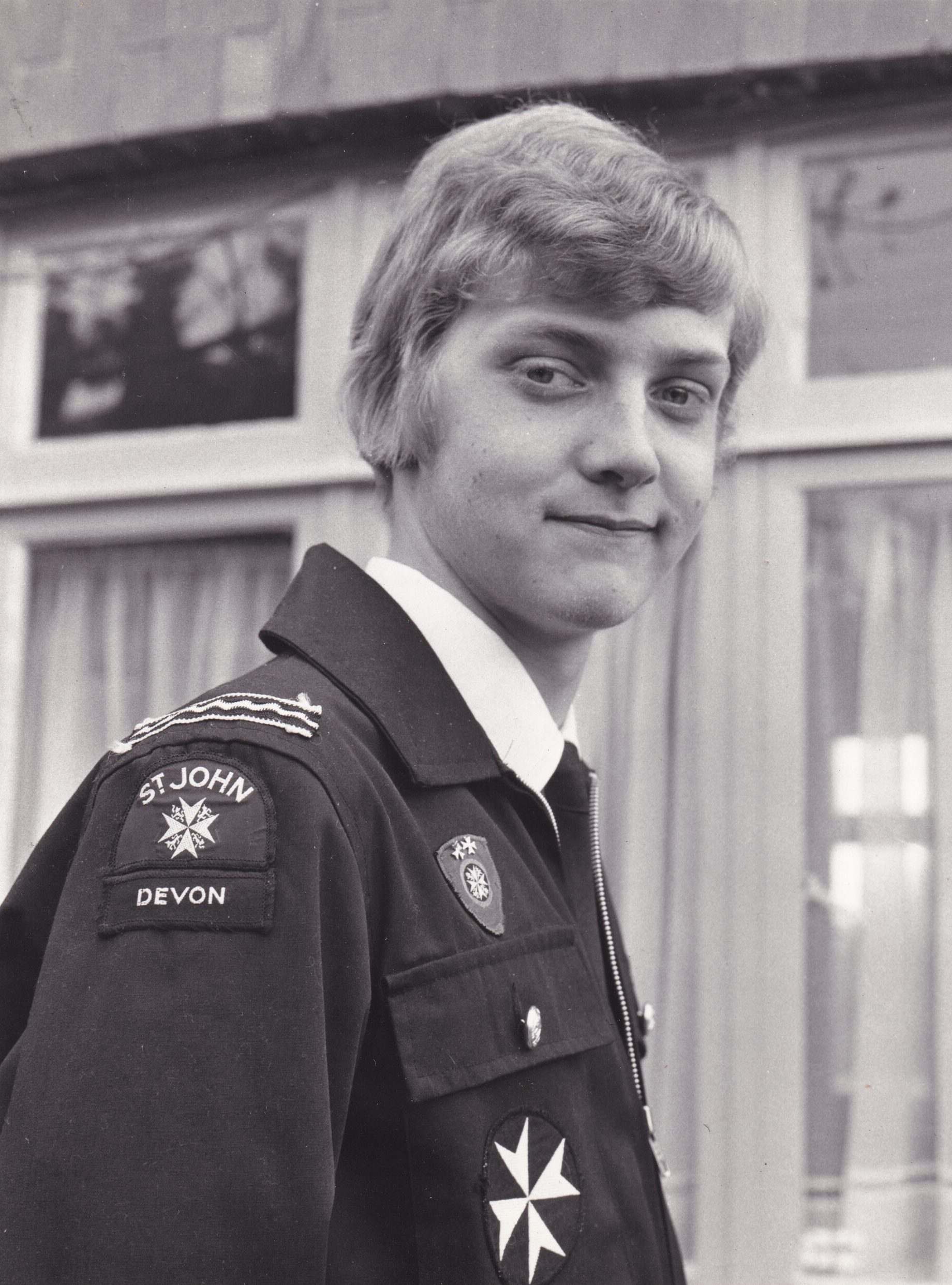 Black and white photograph of a boy wearing a black uniform and looking to the camera over his shoulder. On the sleeve of his jacket are badges that read St John Devon. In the backgrouynd are windows of a house.