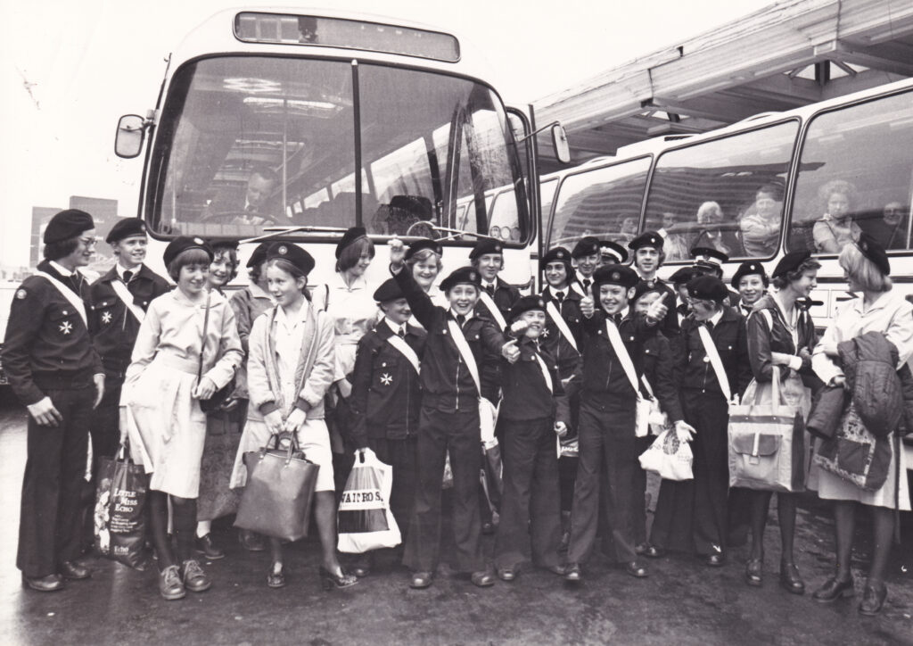 Black and white photograph of a large group of male and female Cadets in uniform standing in front of two coaches. The coach on the right has other young people sitting inside. The Cadets hold bags.