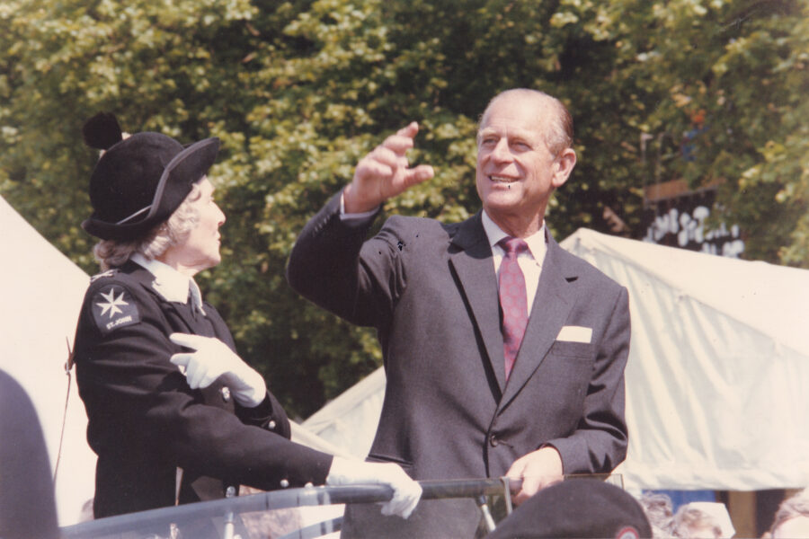 Colour photograph of two people. On the left is the Duke of Edinburgh who is waving. To the right is a woman in St John uniform. She is not facing the camera.
