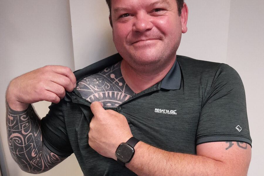 Colour photograph of an adult male in a grey t-shirt. He stands facing the camera and is pulling the neck of his shirt to one side to show a St John Ambulance tattoo on his shoulder.