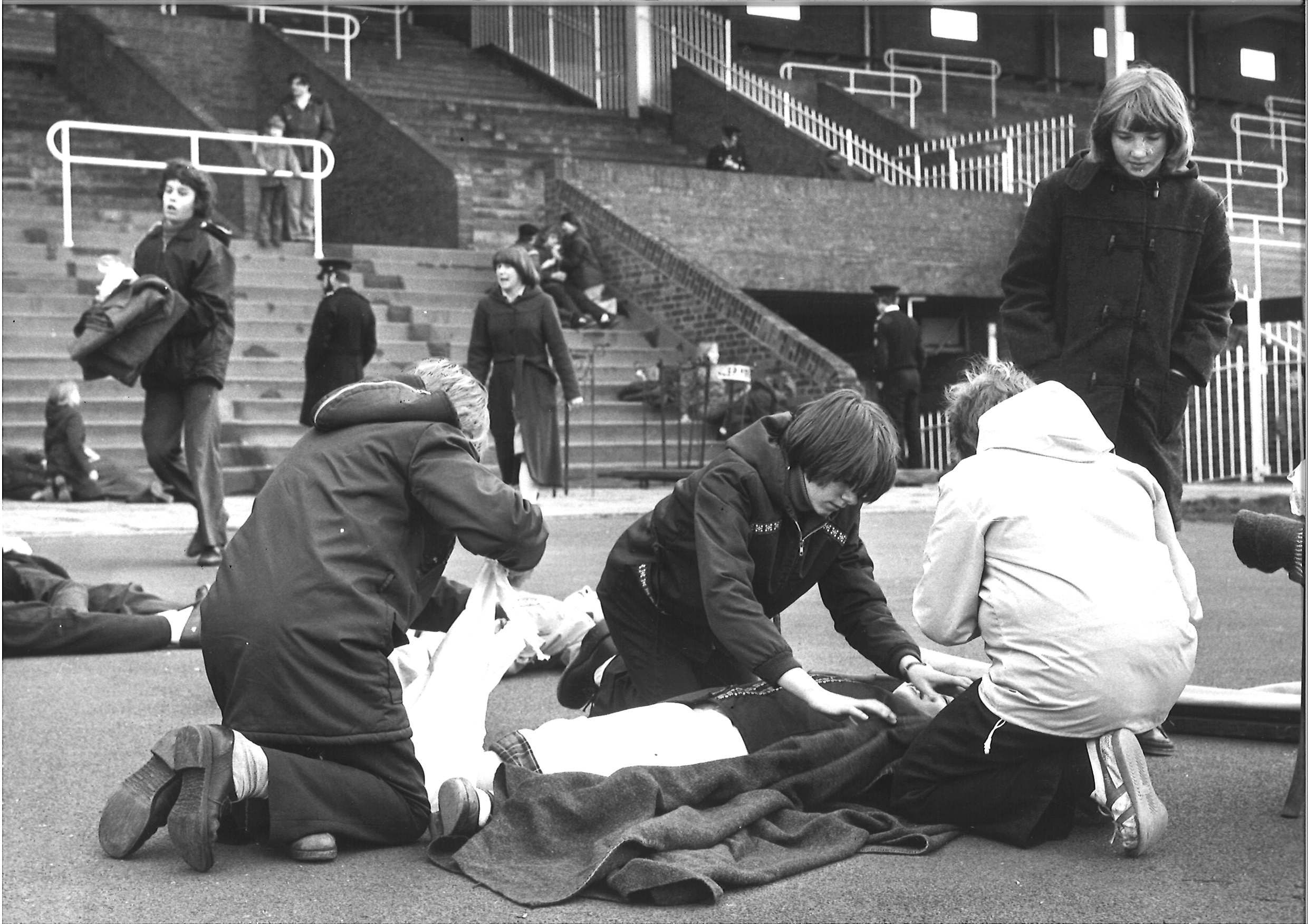 Black and white photograph with three boys in the foreground treating a partially obscured casualty lying on the ground. The boys are wearing non-uniform jackets and a fourth boy stands over them watching in a dark duffle coat. Behind them in the stadium are other small groups carrying out similar exercises.  