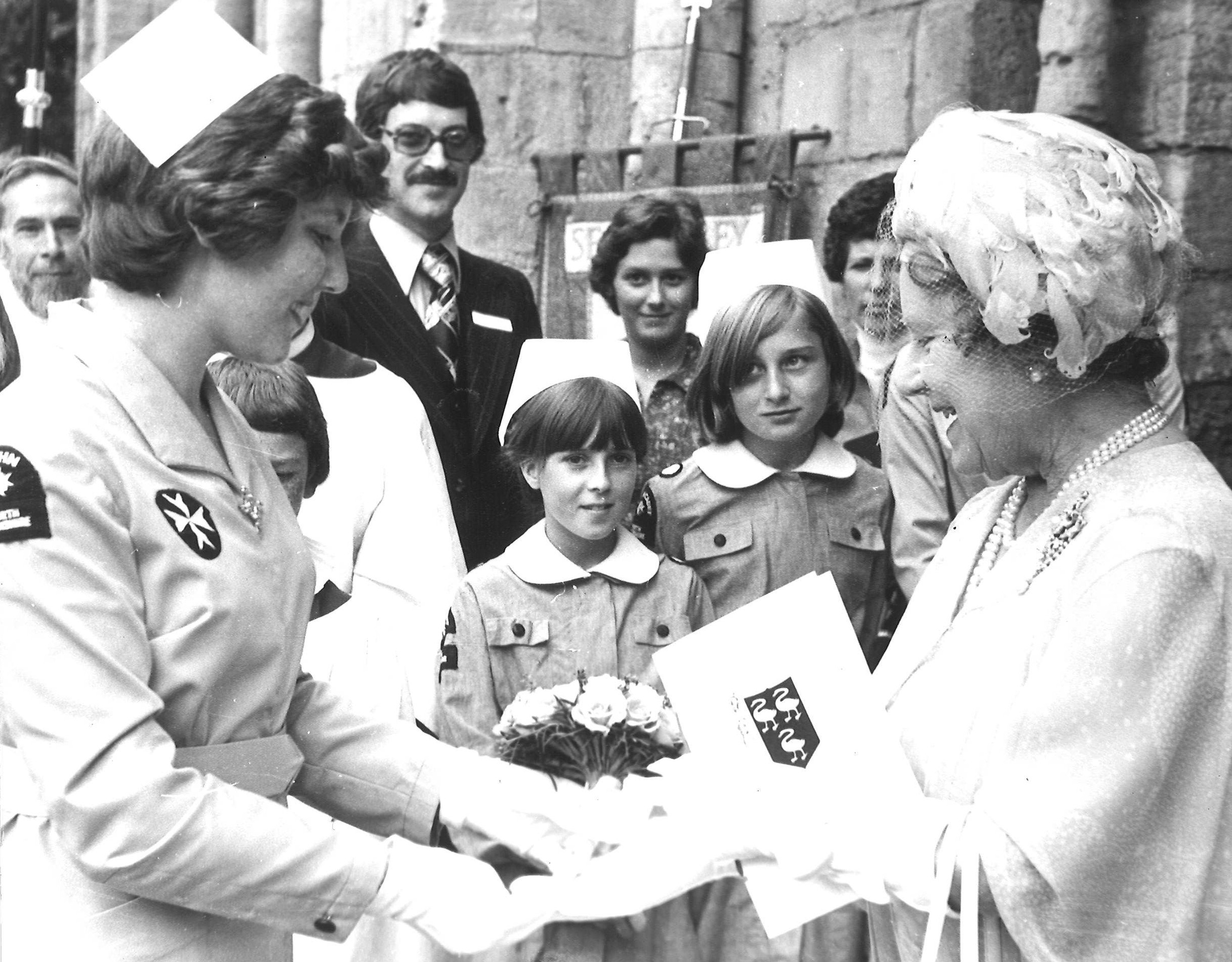 Black and white photograph of Queen Elizabeth II being greeted by a group of young Cadets and adults in uniform. They are presenting the Queen with a small bunch of roses.