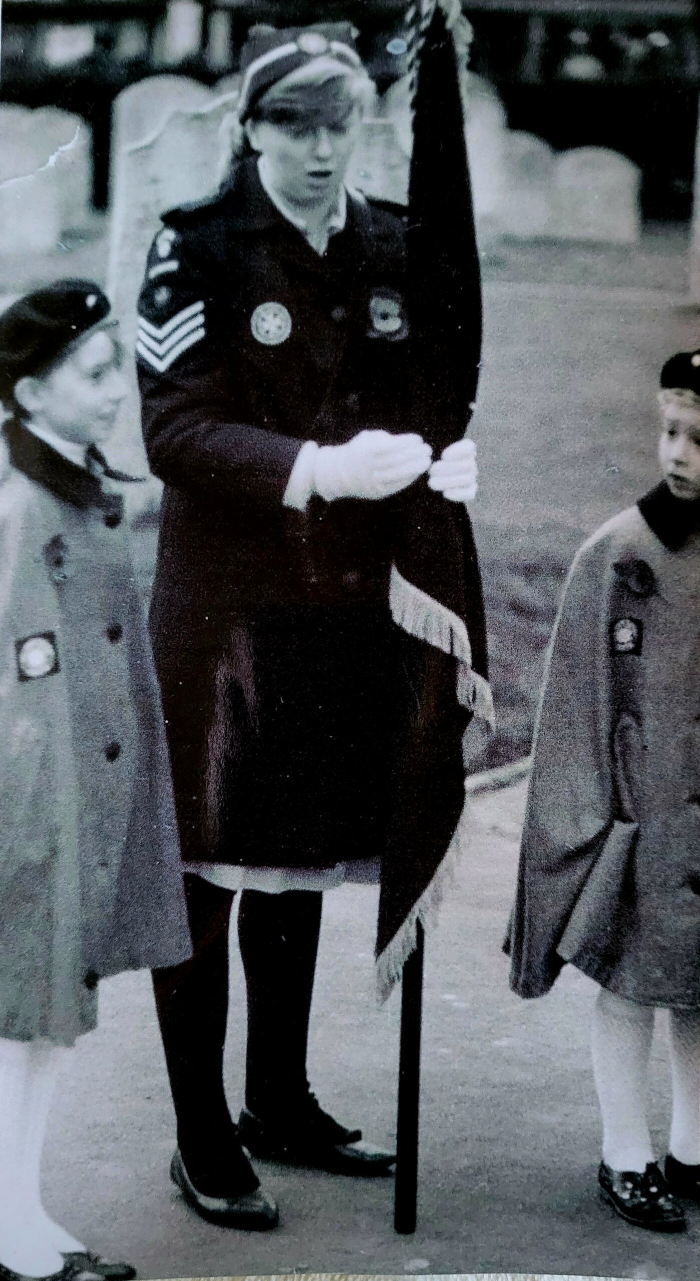 Black and white photograph of three females standing outside. The one is the centre is older and is wearing a dark coloured uniform coat. She is holding a flag in her hands. On either side of her are two younger girls