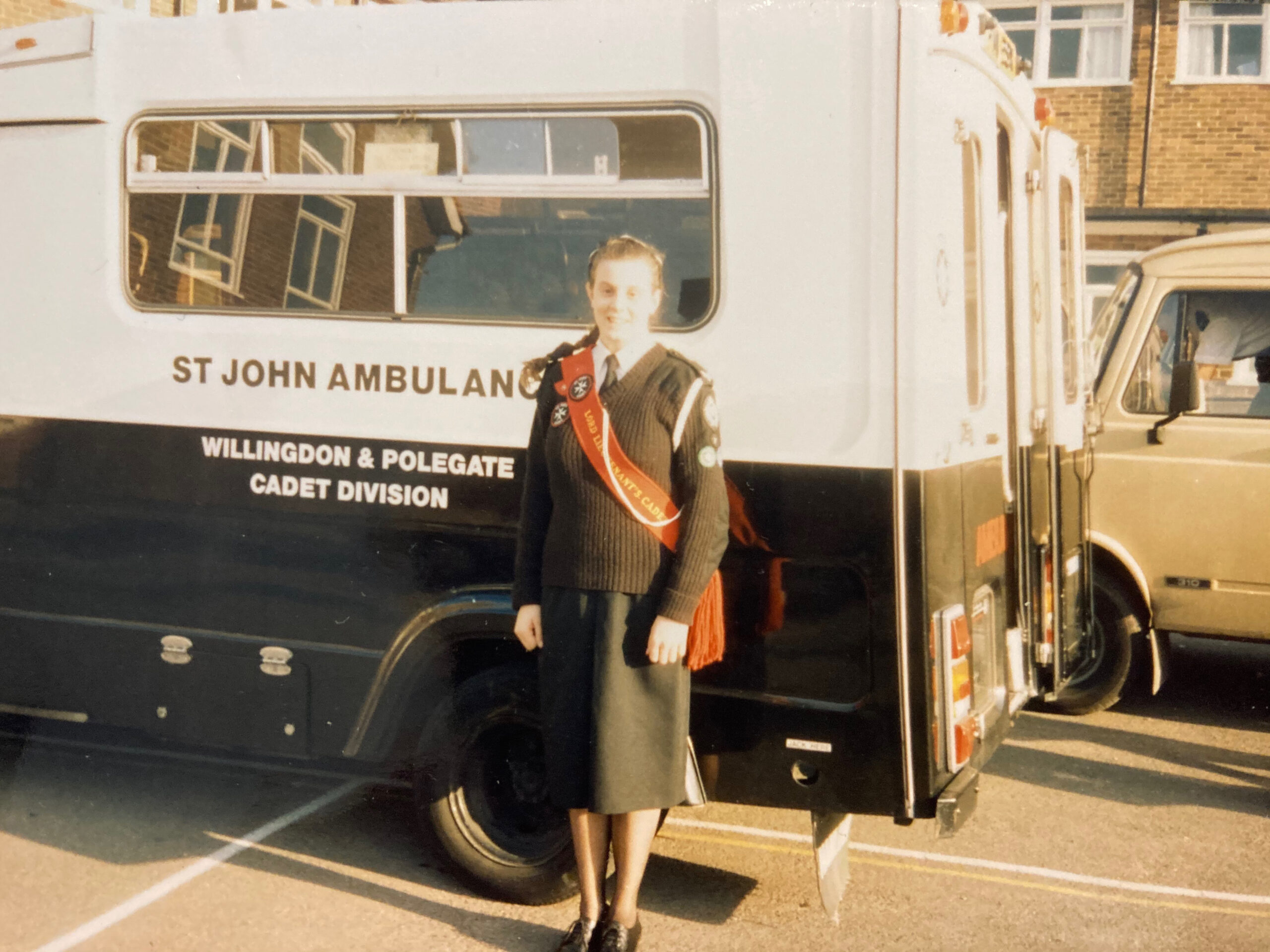 Colour photograph showing a female Cadet standing in front of an ambulance emblazoned with ‘St John Ambulance Willingdon and Polegate Cadet Division’. The Cadet is wearing a white shirt with a black tie