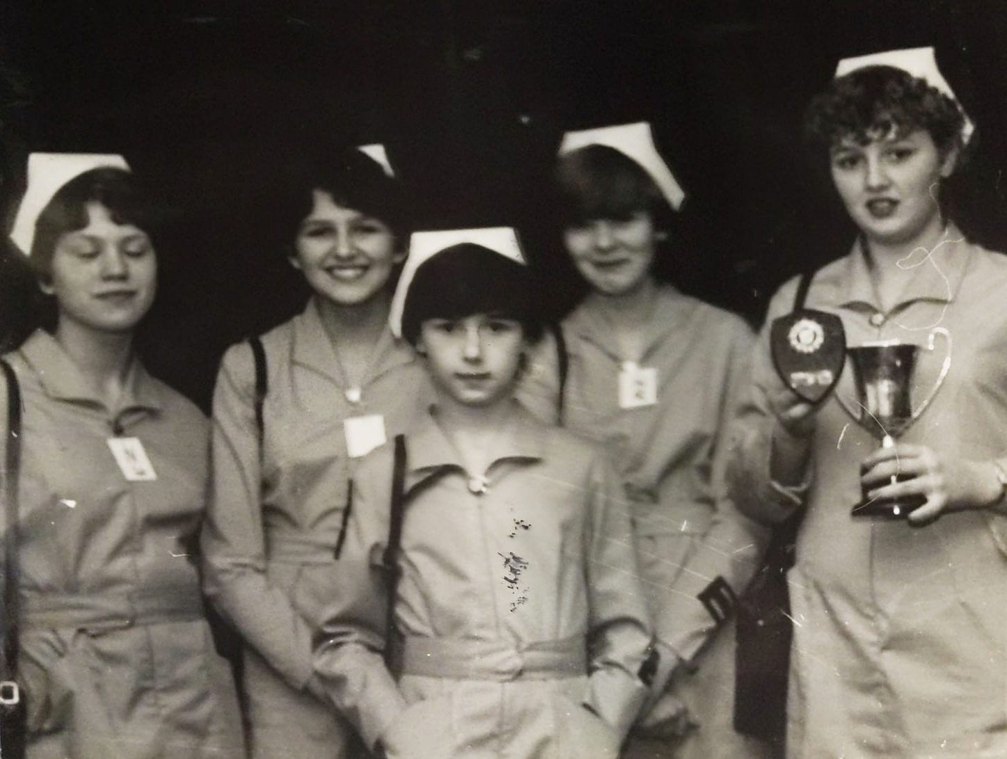 Black and white photograph of five female nursing Cadets in uniform standing in a row. The girl on the end is holding two trophies.