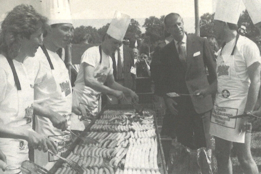 Black and white photograph of four people cooking sausages on a barbeque. They wear chefs hats and aprons. The Duke of Edinburgh is speaking to them.