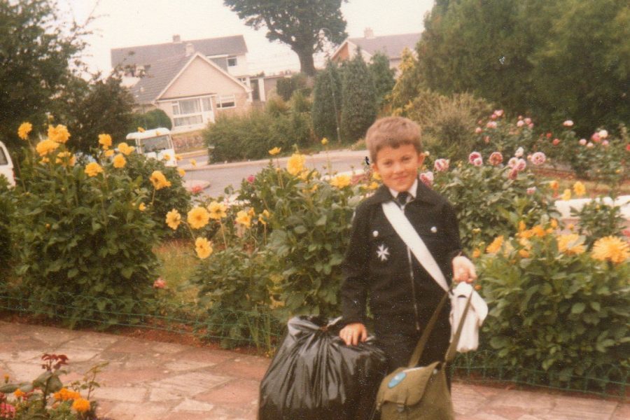 Colour photograph of a young male Cadet in uniform standing on the driveway of a home. Around him are three bags. In the background are rows of flowers.