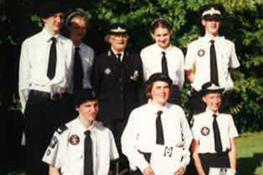 Colour photograph showing seven Cadets. Four are standing at the back with their Cadet Leader in the middle wearing her black St John Number One Uniform consisting of a black jacket and black knee length skirt with a black peaked hat with a white rim and white badge in the centre. Three Cadets are kneeling at the front all holding white certificates and black oval badges. The Cadets all wear black trousers