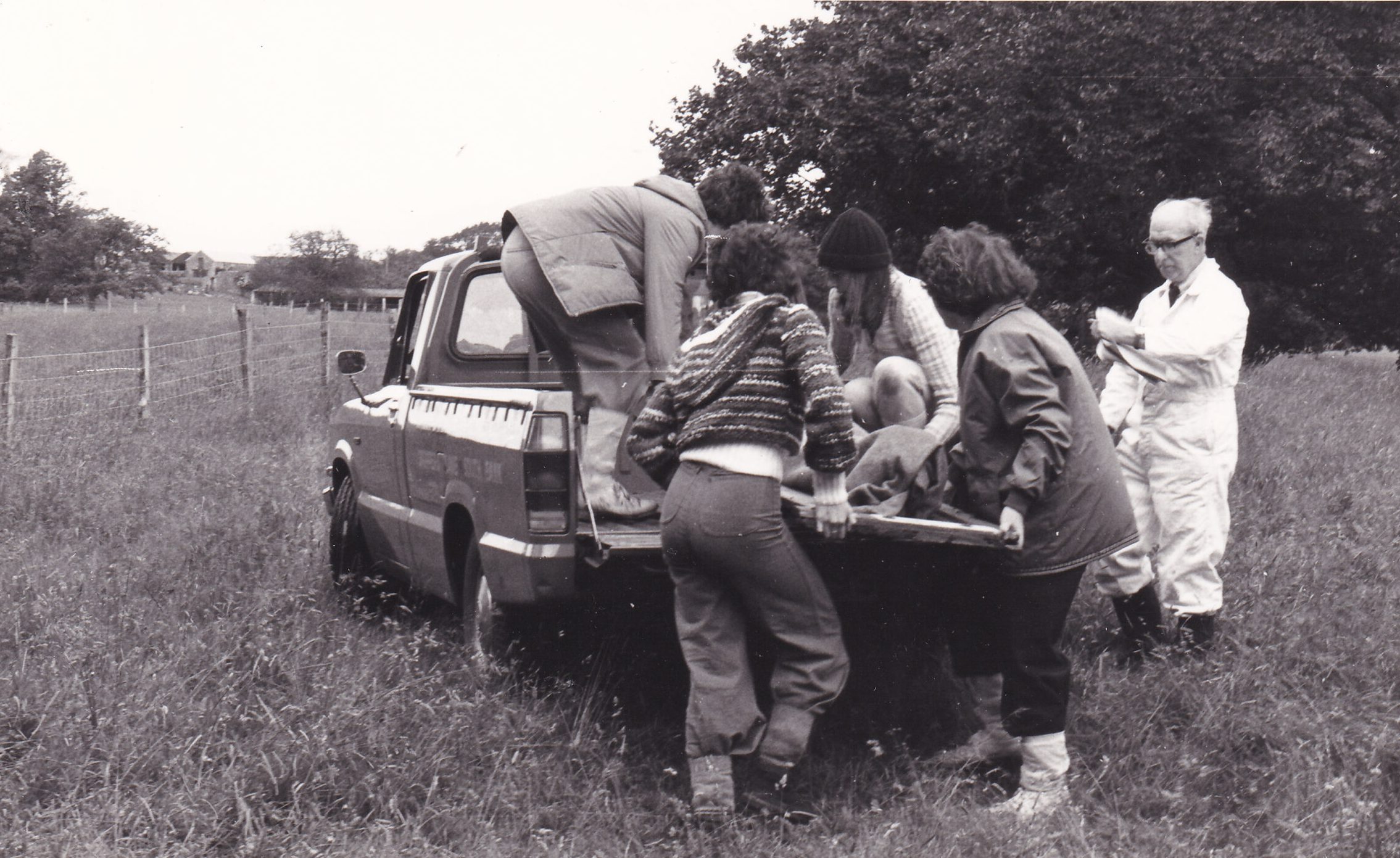 Black and white photograph of four young people lifting a person onto the back of a truck on a stretcher. An adult male looks on and is making notes on a clipboard. The truck is parked in a field.