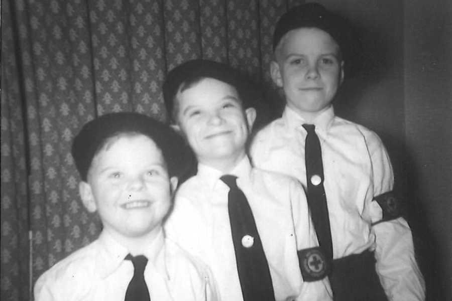 Black and white photograph showing three young white boys posing for a photo in front of dark patterned curtains all smiling at the camera. The boys ascend in height and all are wearing matching uniforms consisting of black berets, white shirts, black ties with a small round badge in the centre, black trousers and a black brassard with white circle on it surrounded by white writing and a red cross in the centre.