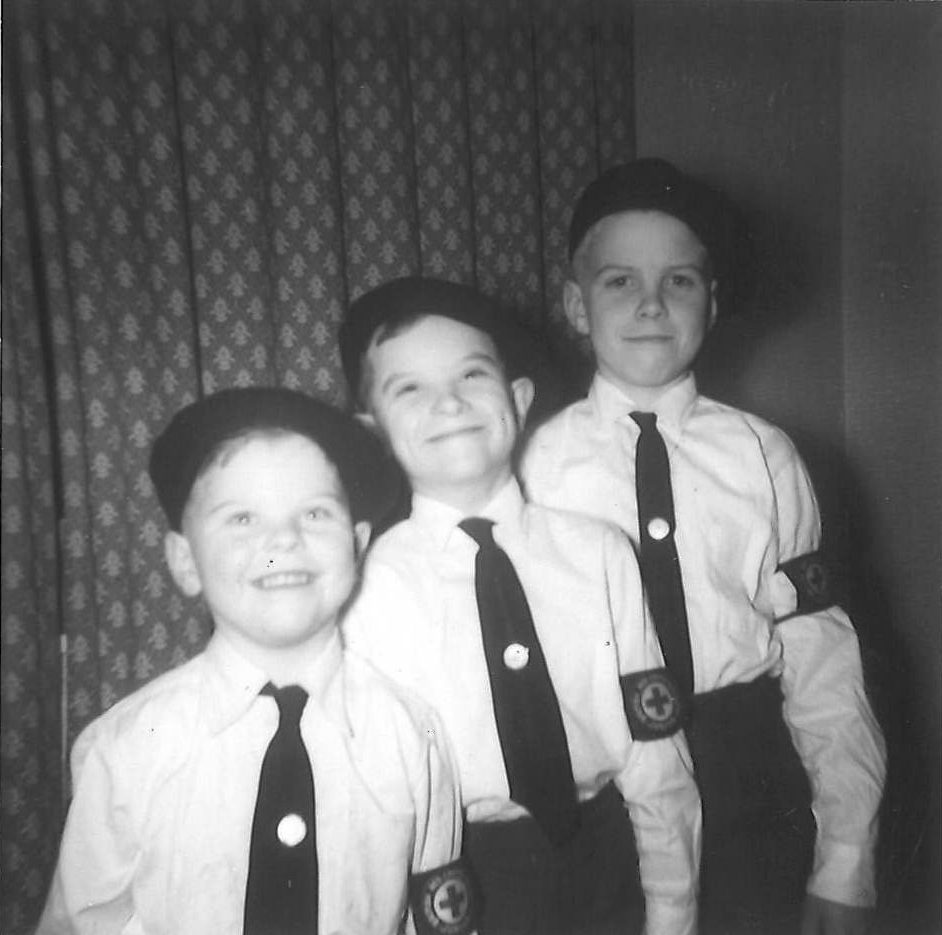 Black and white photograph showing three young white boys posing for a photo in front of dark patterned curtains all smiling at the camera. The boys ascend in height and all are wearing matching uniforms consisting of black berets, white shirts, black ties with a small round badge in the centre, black trousers and a black brassard with white circle on it surrounded by white writing and a red cross in the centre.