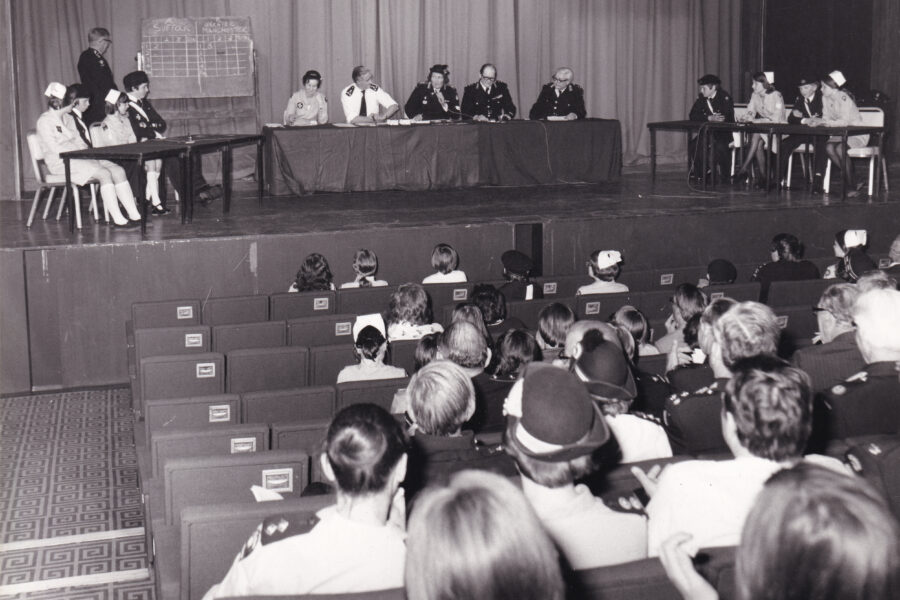 Black and white photograph of a group of people seated in a hall facing a stage. On the stage are three tables. In the centre table sit five adults in uniform. On either side are tables with four young Cadets sitting at each. Behind them is an adult in uniform with a blackboard.