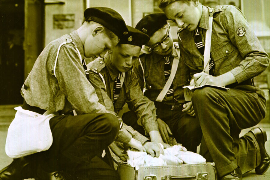 Black and white photograph of four young male Cadets in uniform kneeling around a briefcase that sits open on the ground.