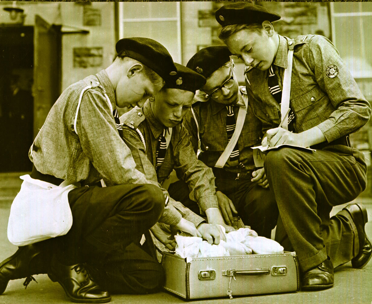 Black and white photograph of four young male Cadets in uniform kneeling around a briefcase that sits open on the ground.