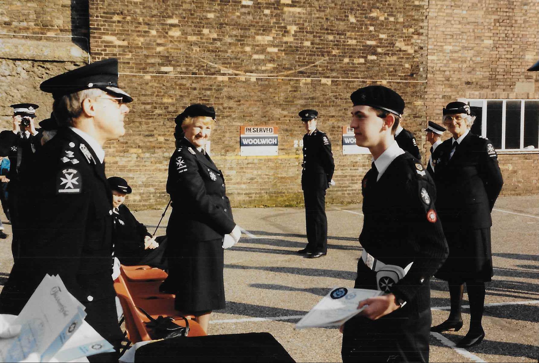 Colour photograph of a male Cadets being met by a group of adults in St John Ambulance uniform. They are standing outside with the brick wall of a building visible behind them.