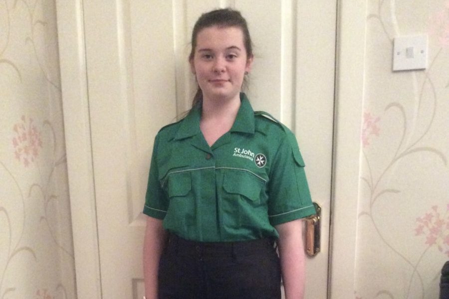Colour photograph of a female Cadet in uniform standing in front of a door. She wears a green St John Ambulance shirt and black trousers.