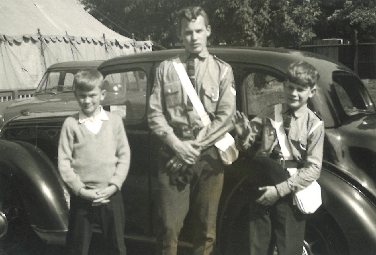 A black and white photo showing three white, smiling boys, in front of a dark car with a white marquee in the background. The middle boy looks older and is much taller than the two on either side of him. The boy to the left wears white shirt, pale V-necked jumper and black trousers. The two boys to the right wear grey shirts with striped ties and grey trousers. Across their bodies they have a white haversack.