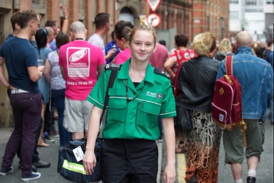 Colour photograph of Chloe Hobson in green and black St John uniform, carrying First Aid kit, with a crowd of people in the background