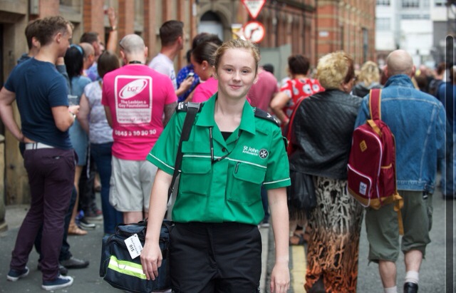 Colour photograph of Chloe Hobson in green and black St John uniform, carrying First Aid kit, with a crowd of people in the background