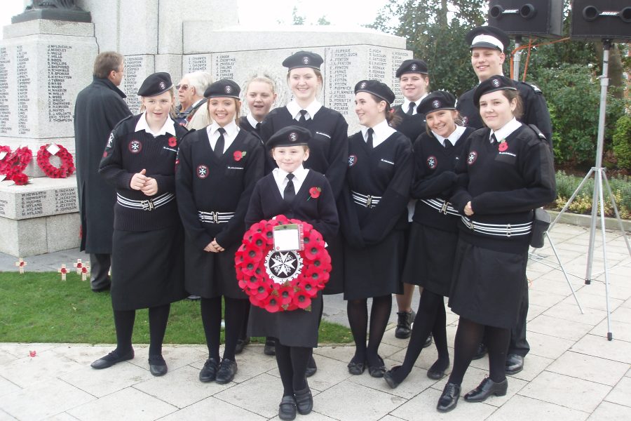 Colour photograph of 8 Cadets in black and white St John uniform posing for a photograph on Remembrance Sunday, Chloe Hobson holds a wreath of poppies in the front row.