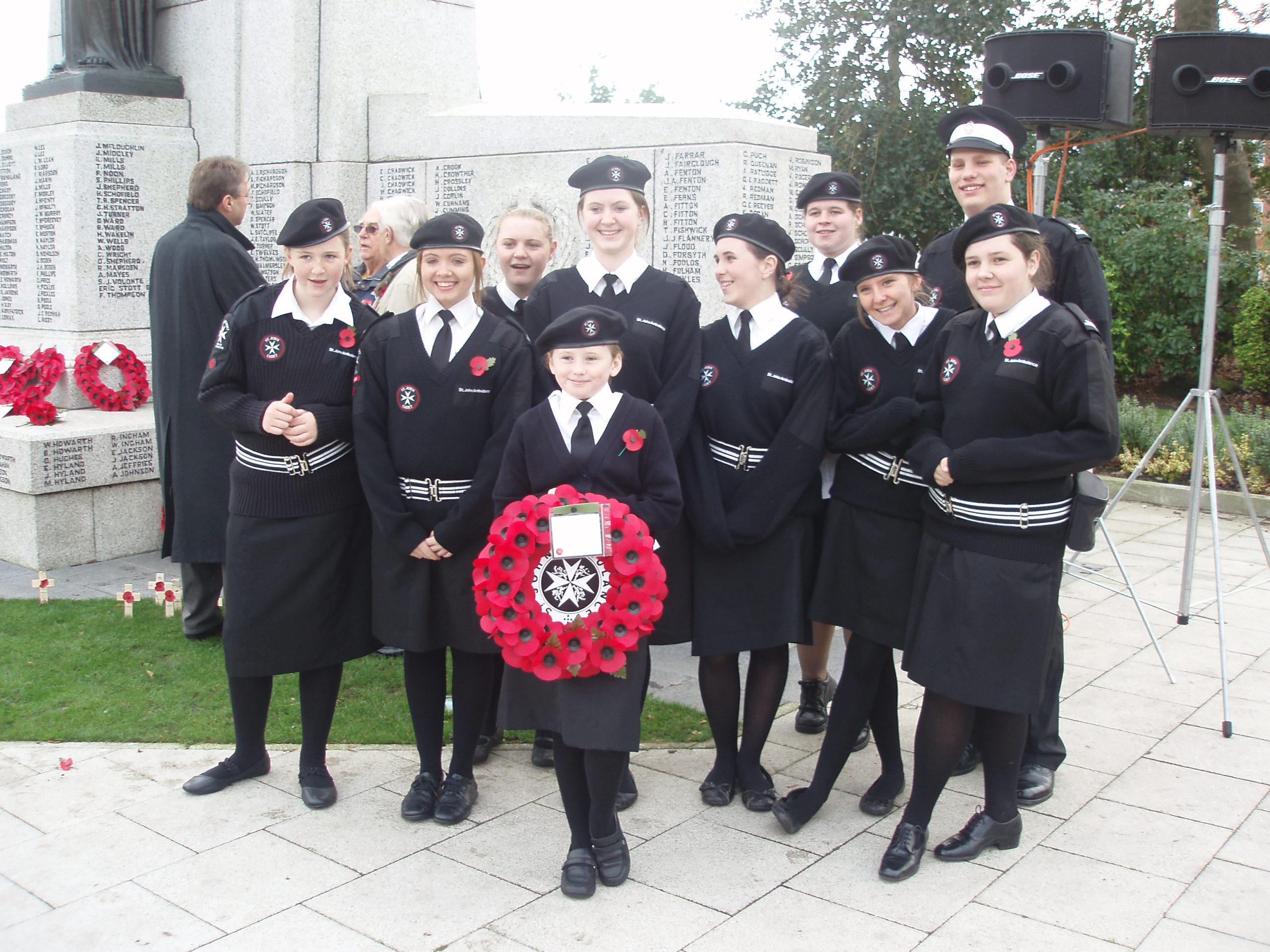 Colour photograph of 8 Cadets in black and white St John uniform posing for a photograph on Remembrance Sunday, Chloe Hobson holds a wreath of poppies in the front row.