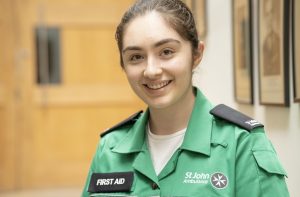 Colour photograph of a young woman in green and black St John uniform standing in a corridor and smiling at the camera