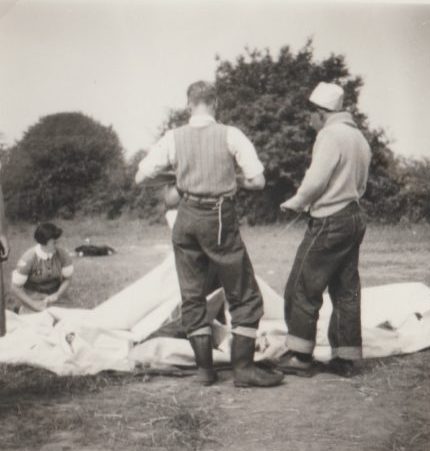 Black and white polaroid showing two men or boys, viewed from behind, standing in front of some partially constructed tents in a field, a female Cadet in uniform sat on the ground beyond the tents