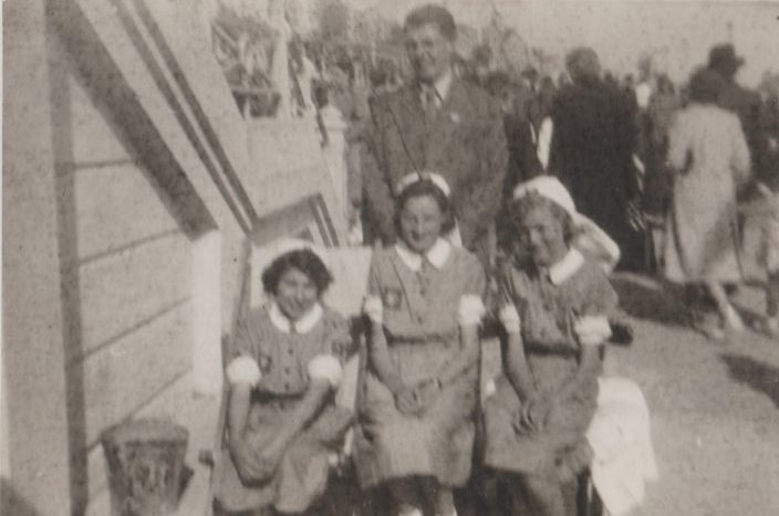 Blurred black and white photograph showing three Nursing Cadets seated in deckchairs at the bottom of a staircase onto a busy promenade. Behind them stands a man in a suit. The Cadets all wear grey knee length dresses with white nursing caps and white cuffs at their elbows.  